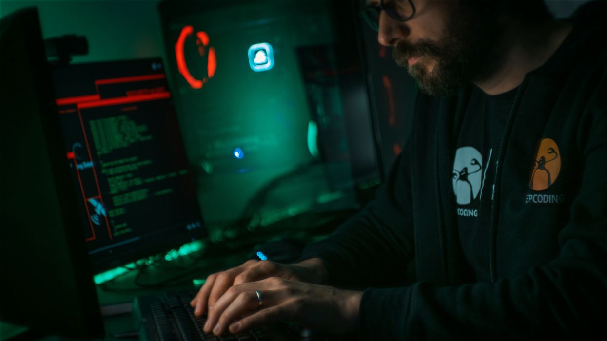a bearded man sits typing on a keyboard in a darkened room with green computer code pictured on-screen