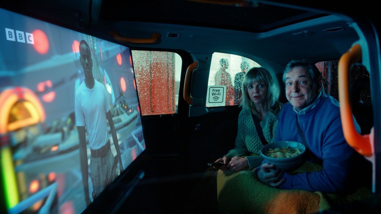 a couple sit in the back of a black cab with crisps and a tv remote watching television on freely