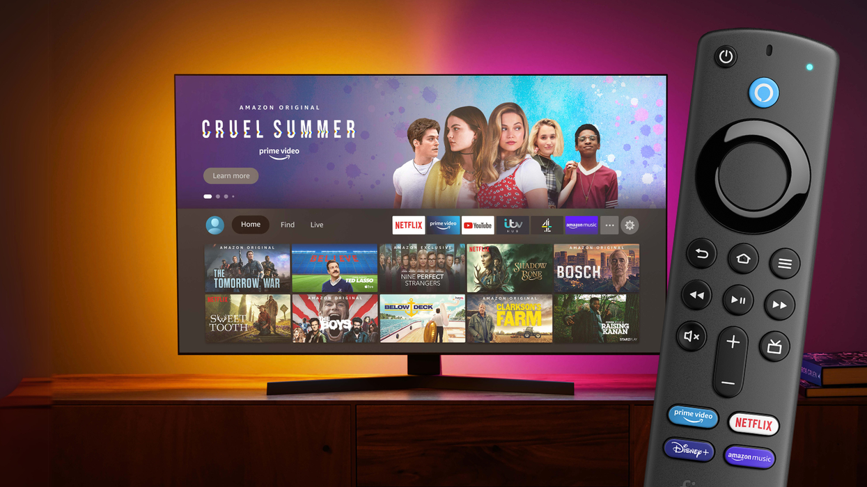 a fire tv stick remote is pictured in the foreground while a living room with a flatscreen tv with the fire tv os on-screen is pictured in the background