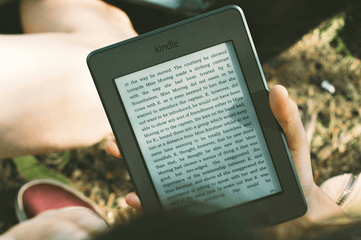 a hand holds a kindle with grass pictured in the background on a summers day