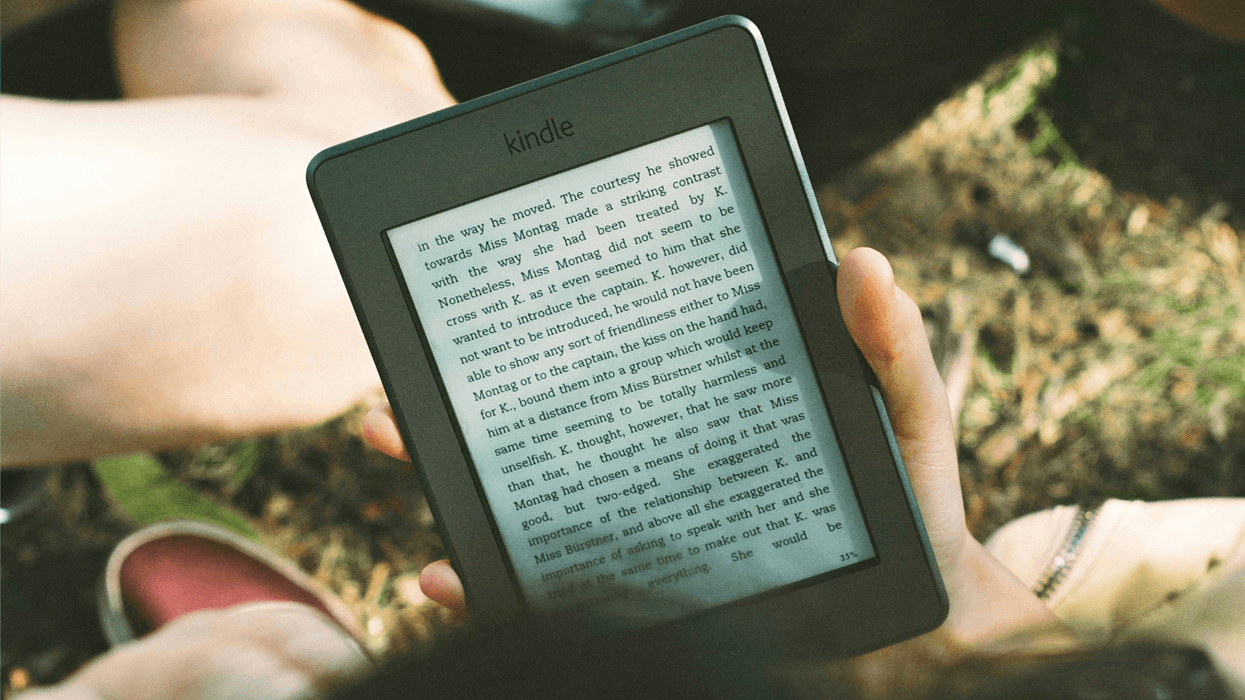 a hand holds a kindle with grass pictured in the background on a summers day