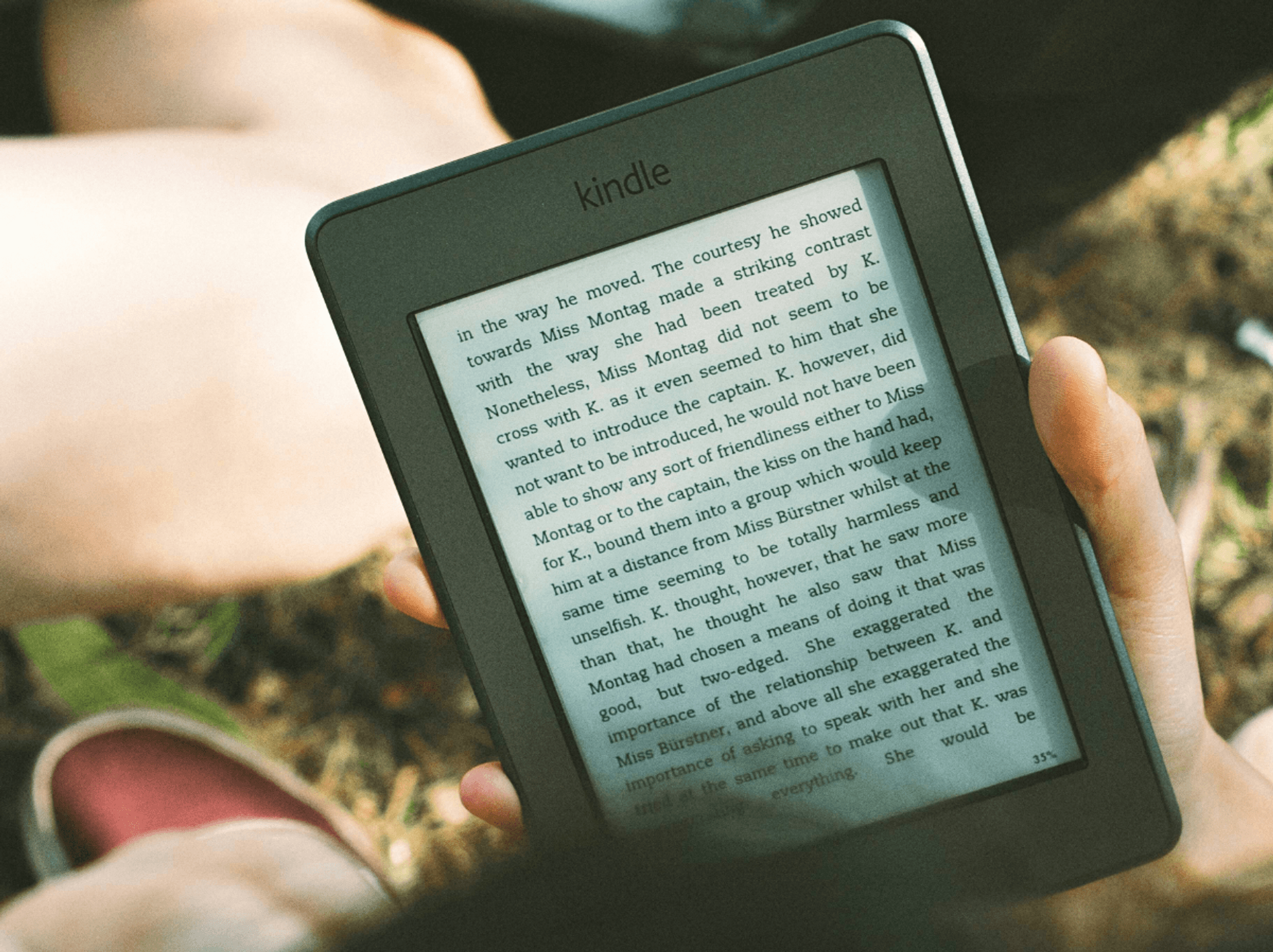 a hand holds a kindle with grass pictured in the background on a summers day