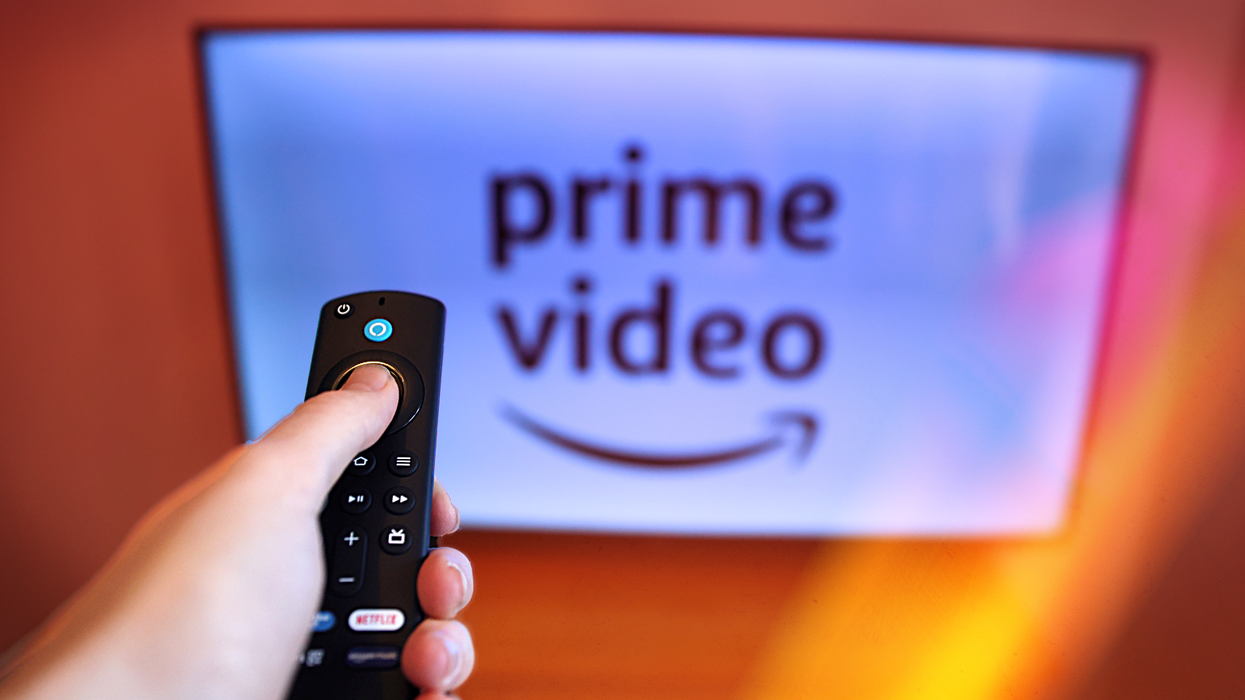 a hand holds a remote control in front of a television with the prime video logo on-screen