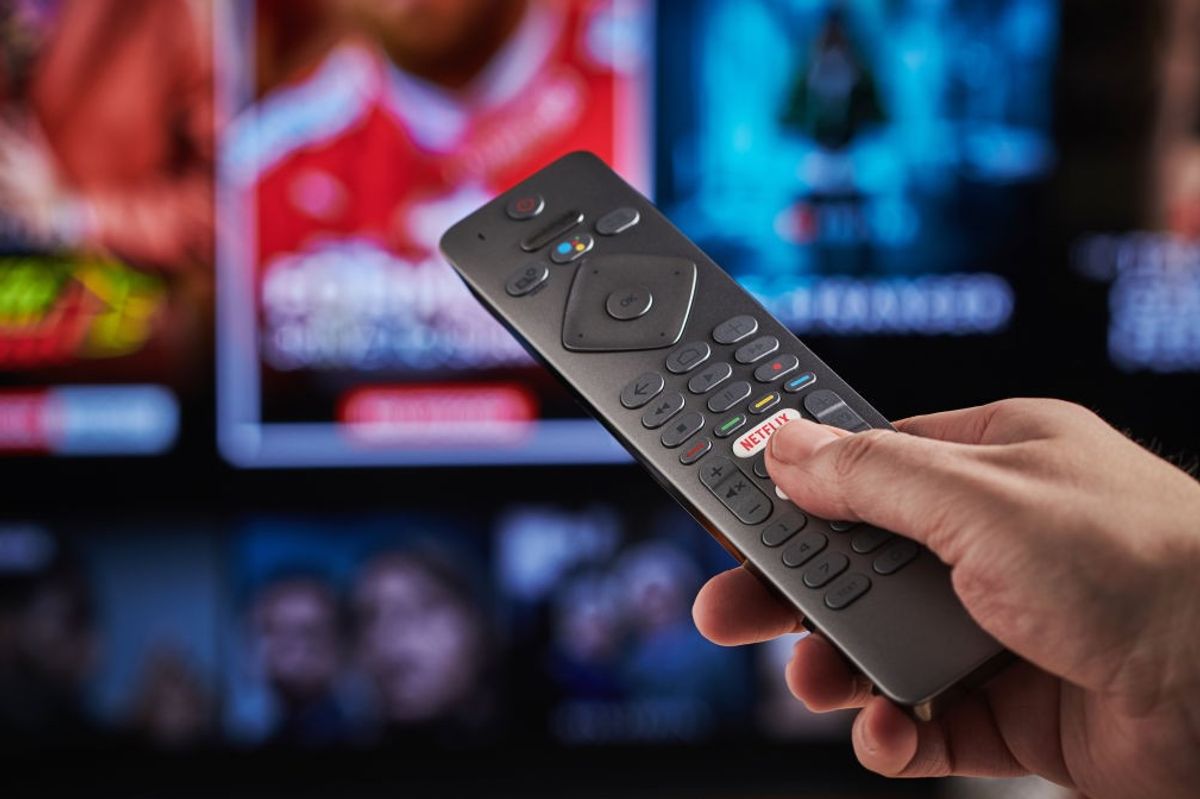 a hand holds a tv remote control with a blurred background of tv show previews in the background  