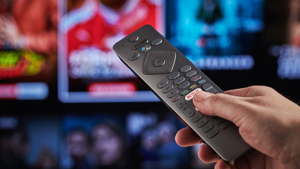 a hand holds a tv remote control with a blurred background of tv show previews in the background