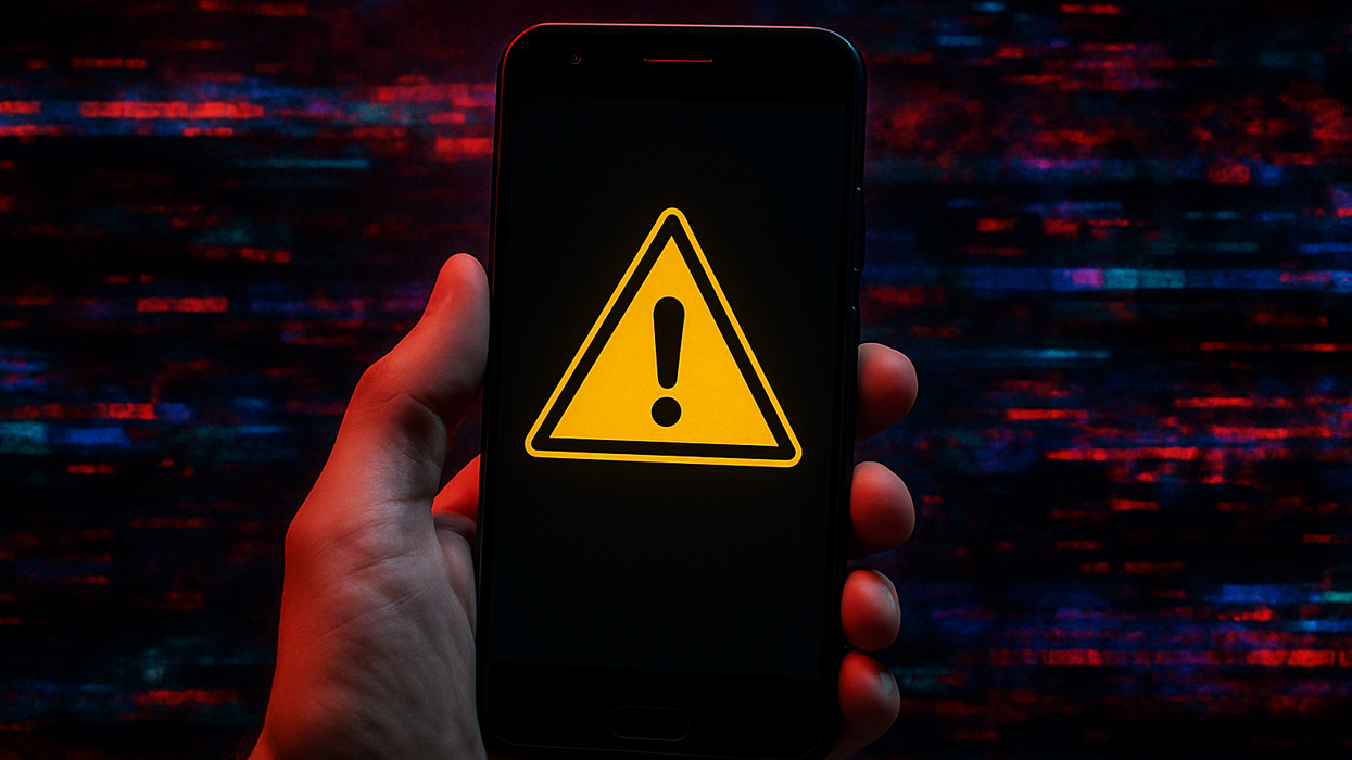 a hand holds an android phone with a warning triangle on-screen, computer glitches can be seen in the background