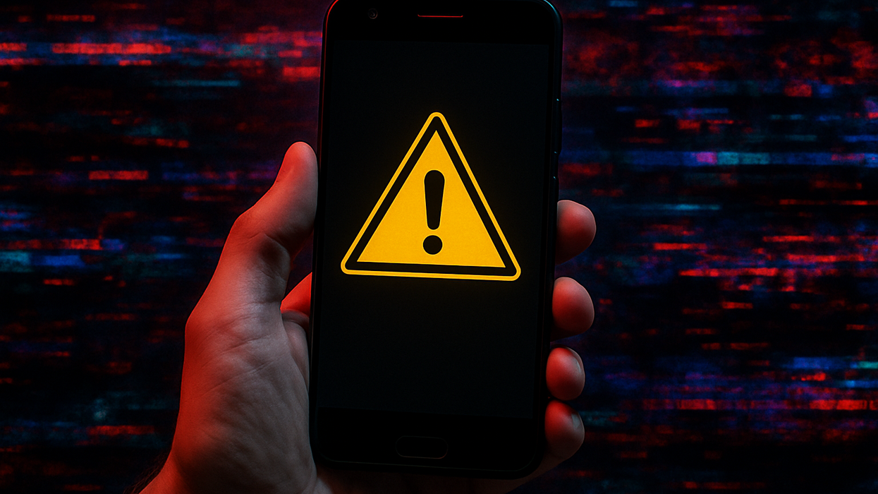 a hand holds an android phone with a warning triangle on-screen, computer glitches can be seen in the background