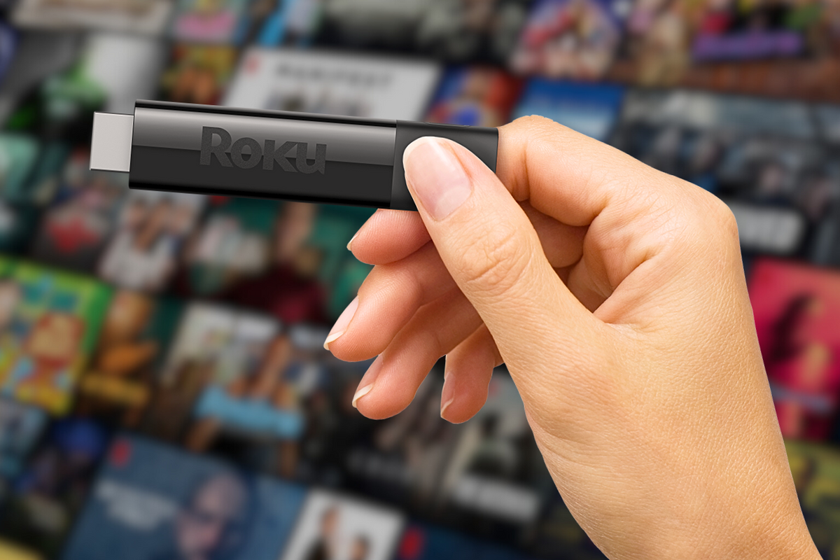 a hand is pictured holding a roku streaming dongle with a blurred artwork for streaming shows in the background 