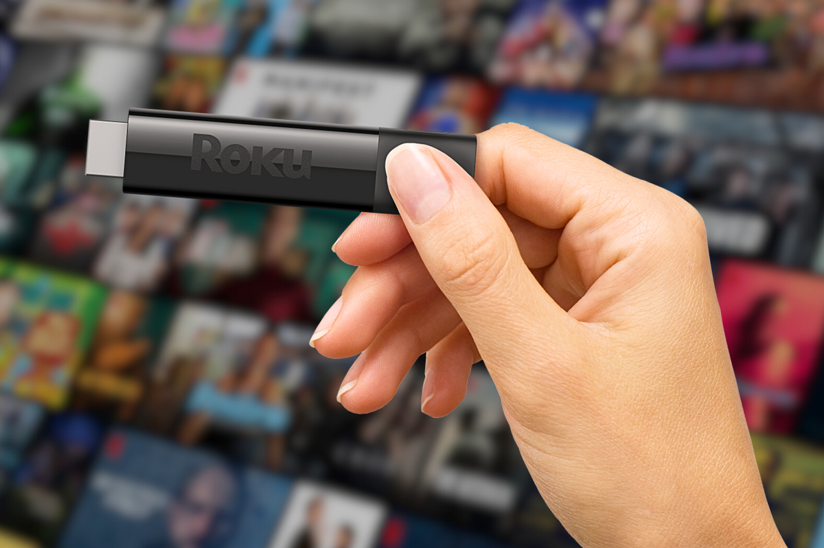 a hand is pictured holding a roku streaming dongle with a blurred artwork for streaming shows in the background 