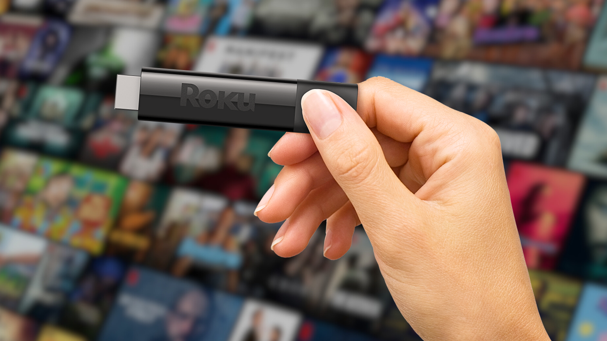 a hand is pictured holding a roku streaming dongle with a blurred artwork for streaming shows in the background