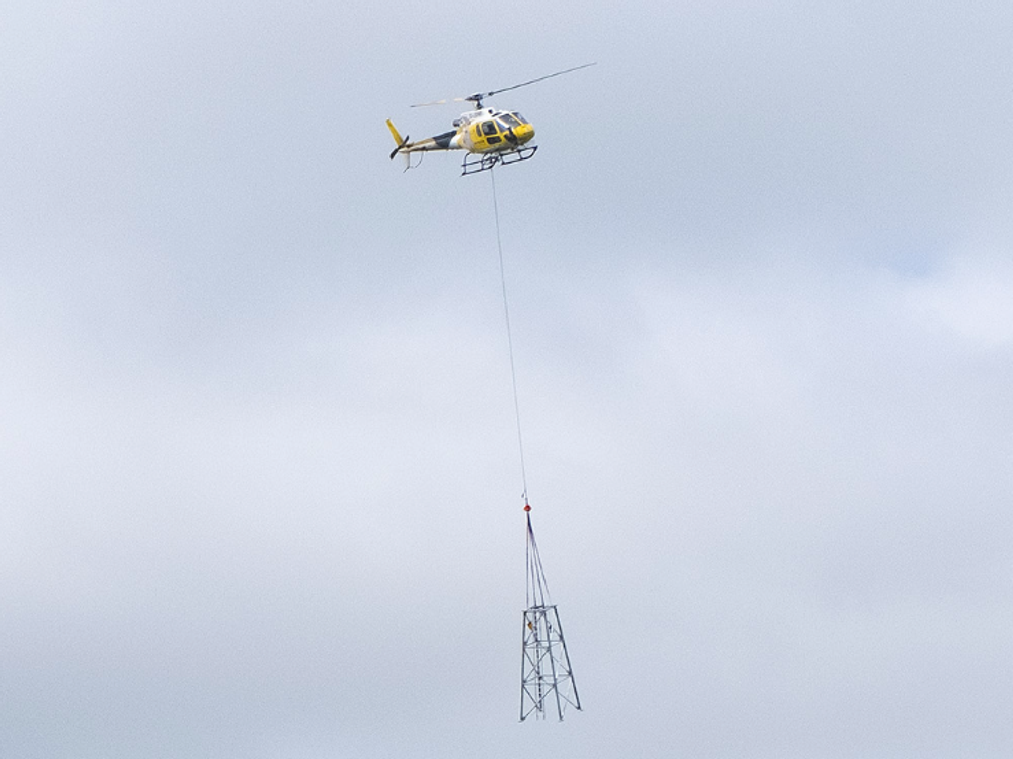 a helicopter is pictured with a piece of a mobile data masts suspended on a wire