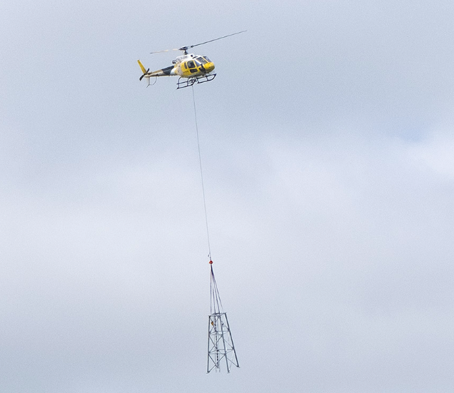 a helicopter is pictured with a piece of a mobile data masts suspended on a wire