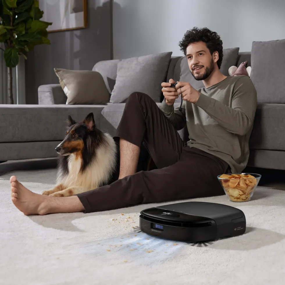 a man sits on the floor next to his dog while the eufy robot vacuum cleanign around him