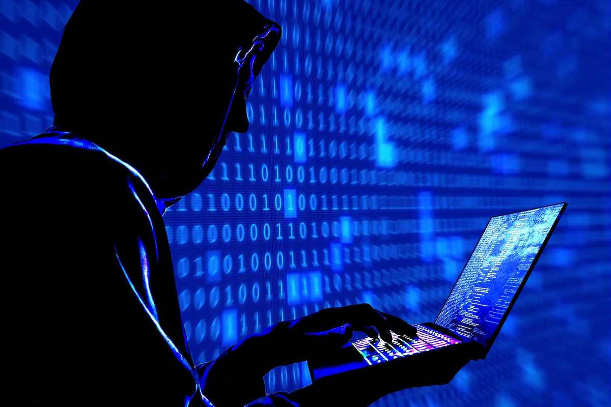 a man with his hood pulled up is pictured typing on a laptop with a blue background of code