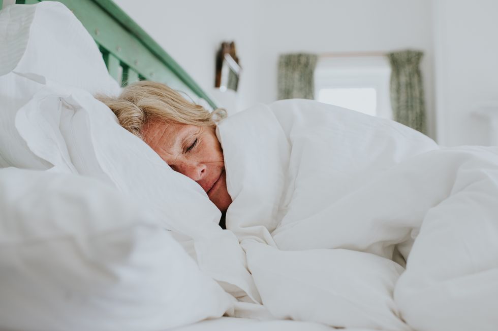 A mature woman cosies up in a clean, white duvet in a bright and airy bedroom