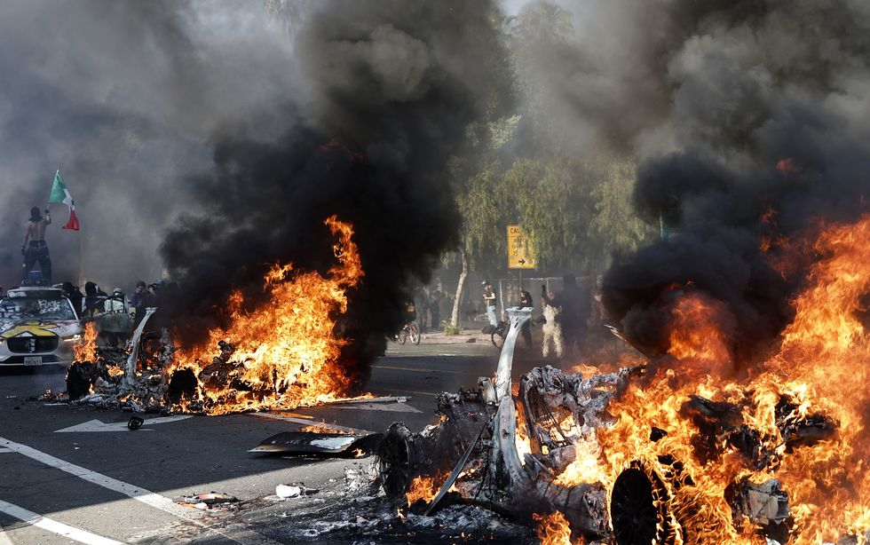 A protester holds up a Mexican flag as burning Waymo cars line the street in Los Angeles, California