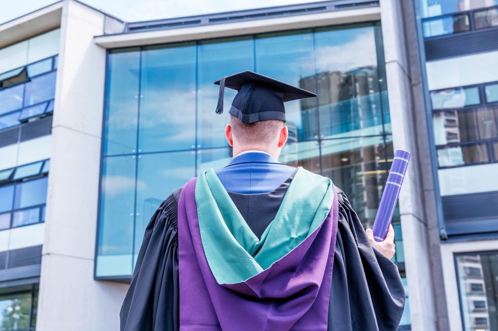 A rear view of a university graduate in a graduation gown holding a diploma in his hand outside the academic building