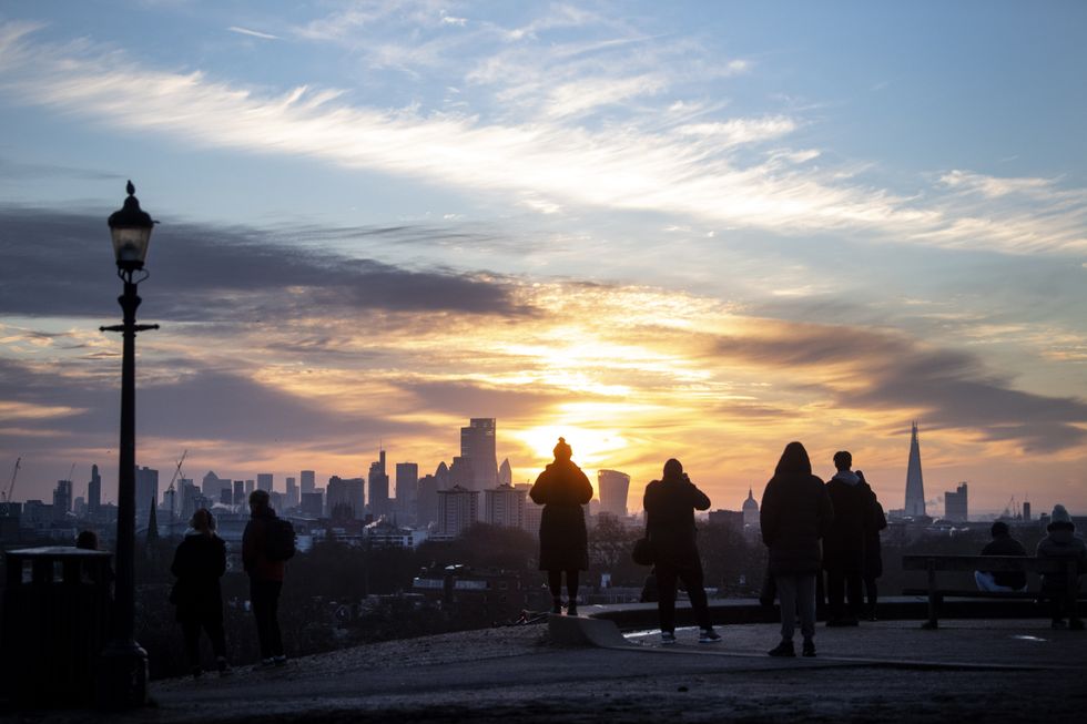 A small crowd of people watch as the sun rises over the city skyline from Primrose Hill, London.