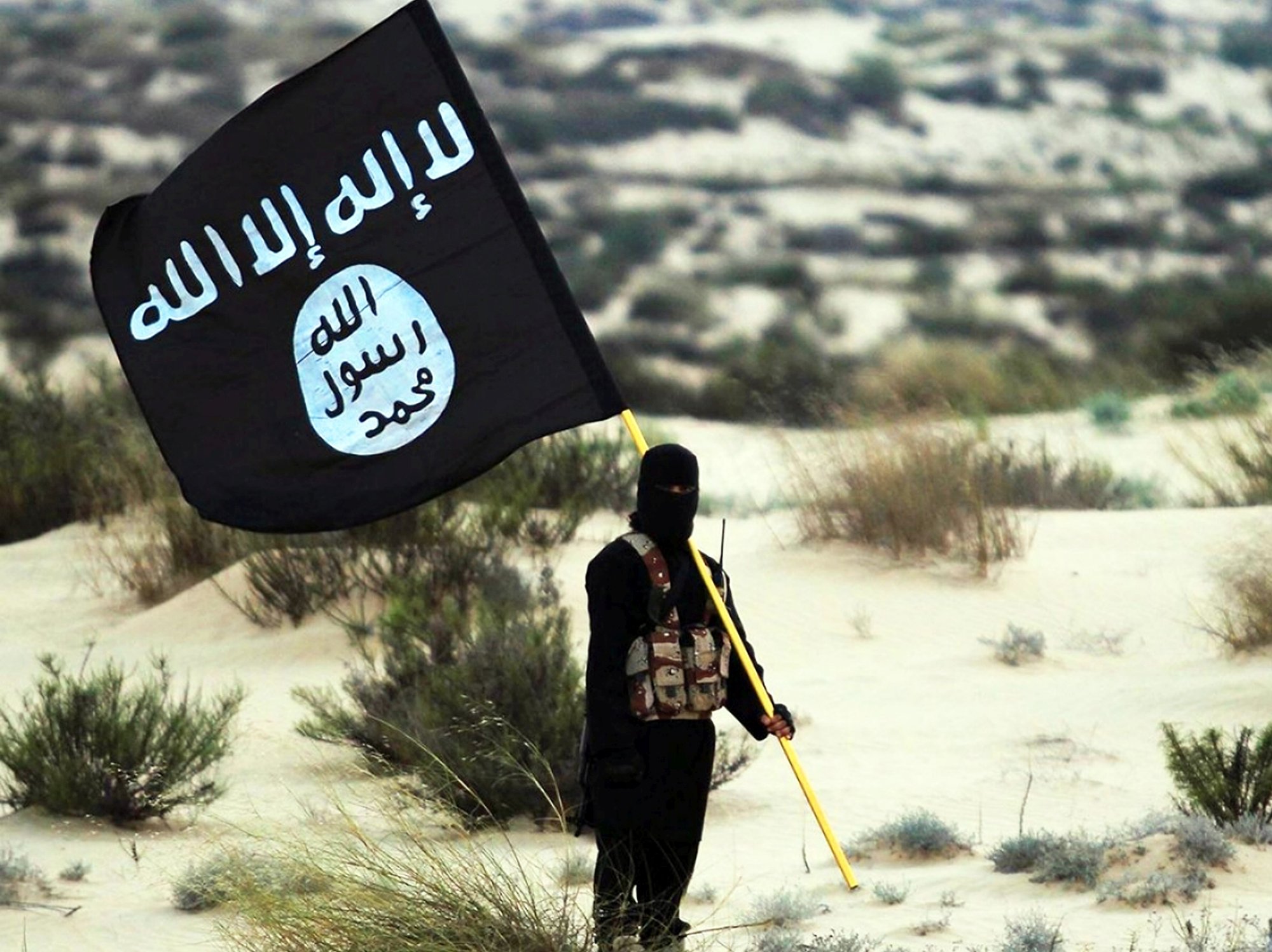 A stock image of a masked Islamic State soldier poses holding the ISIL banner somewhere in the deserts of Iraq or Syria