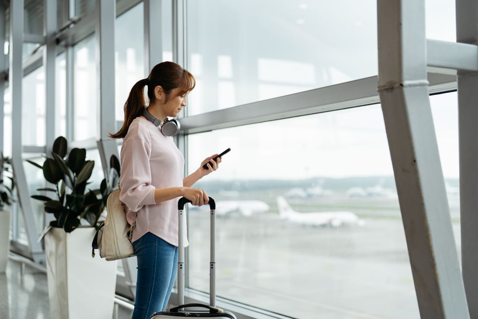 a woman checks her smartphone while holding a pullalong suitcase in the airport 