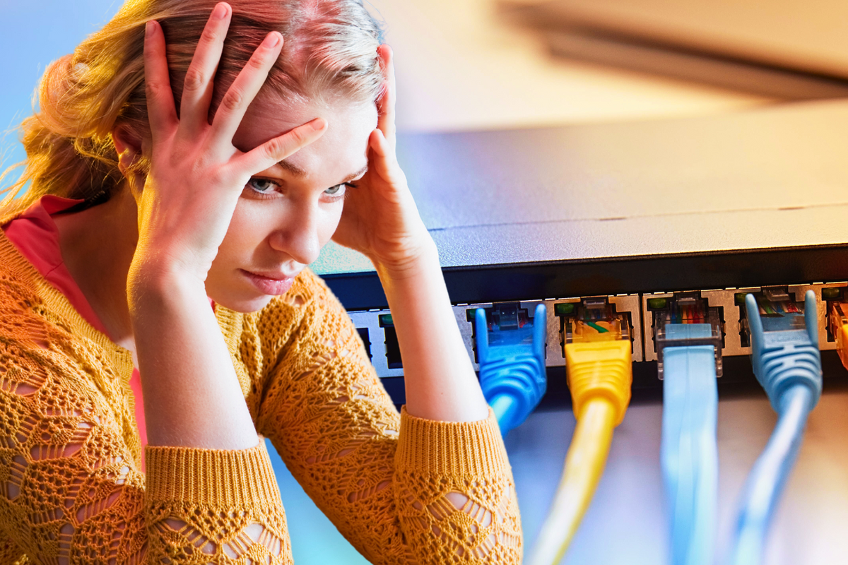 a woman holds head in her hands while a broadband router with ethernet cables connected in pictured in the background  