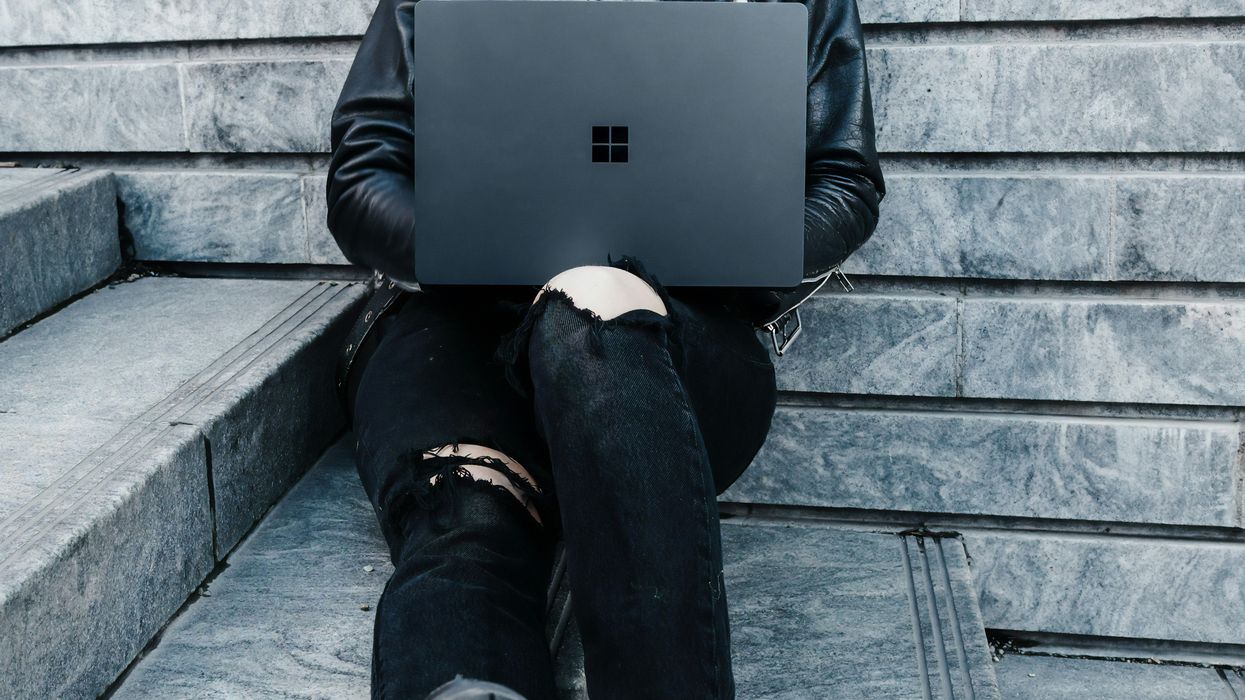 a woman in a leather jacket and jeans sits with a surface laptop on her knees