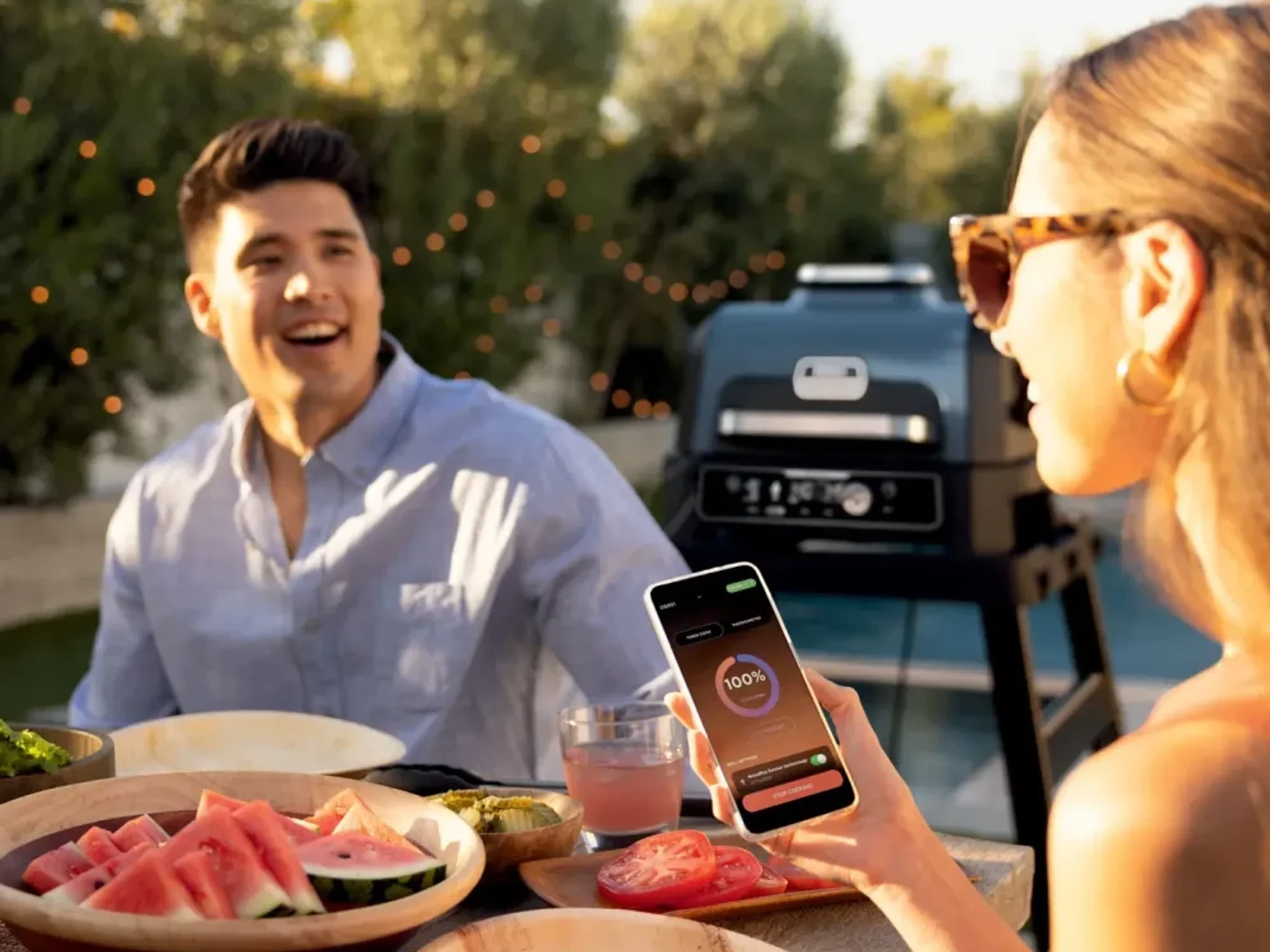 a woman looks at a progress timer on her smartphone to check when the woodfire bbq has finished cooking