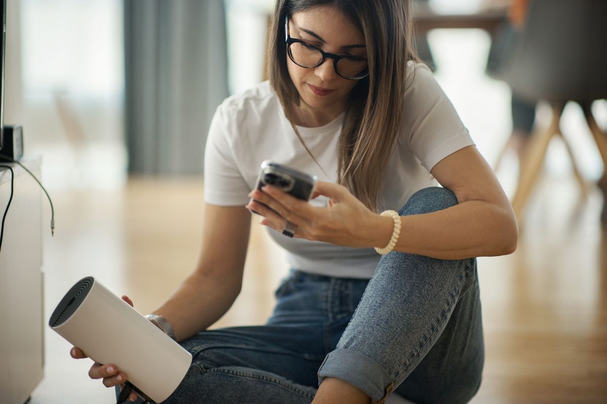 a woman runs a broadband speed test on her phone while holding her wi-fi router sat on the floor