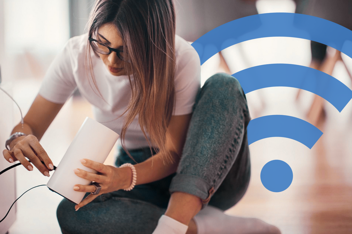 a woman sits cross-legged on the floor and plugs an ethernet cable into the back of her wi-fi router to get internet running 
