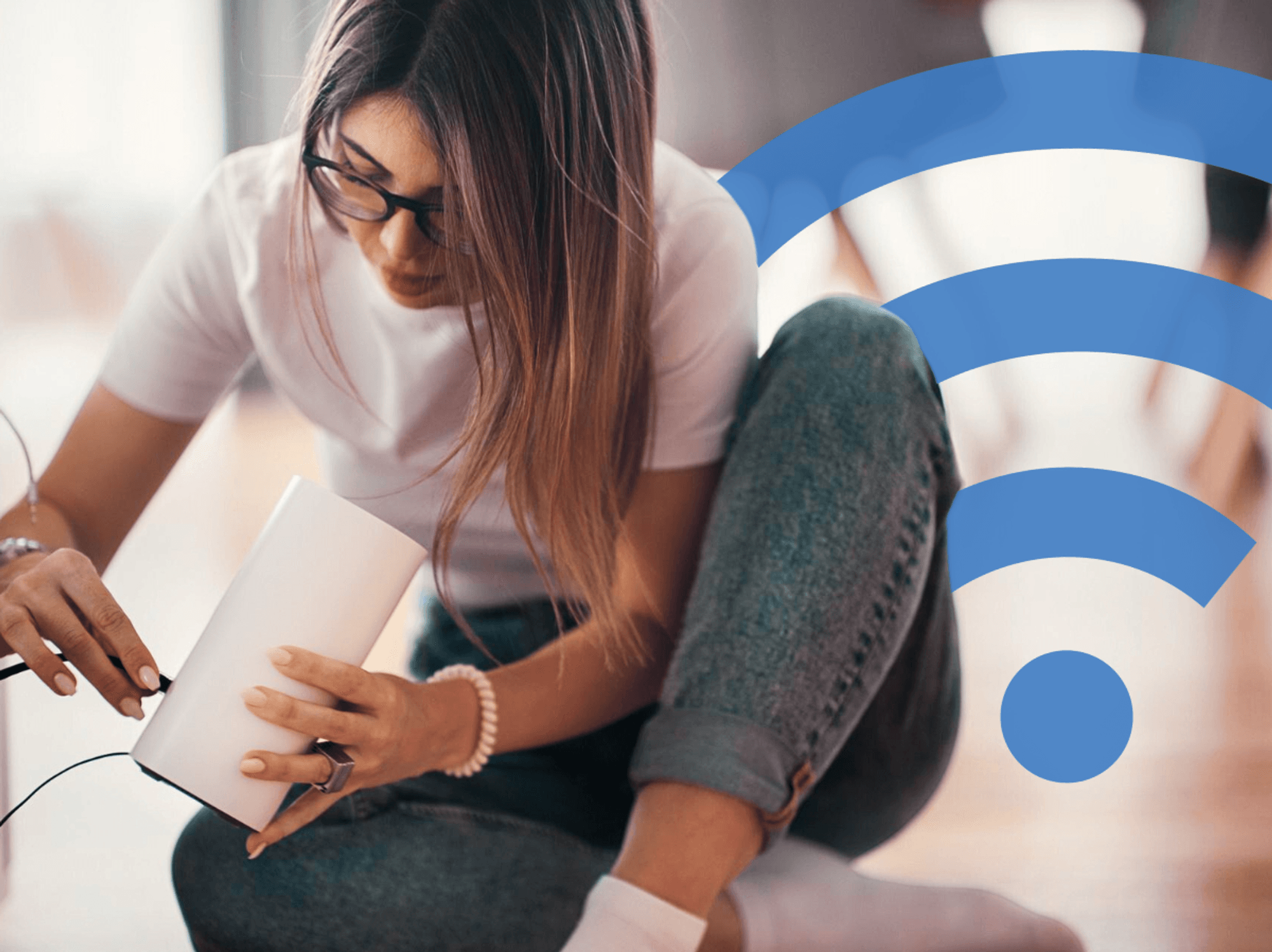 a woman sits cross-legged on the floor and plugs an ethernet cable into the back of her wi-fi router to get internet running