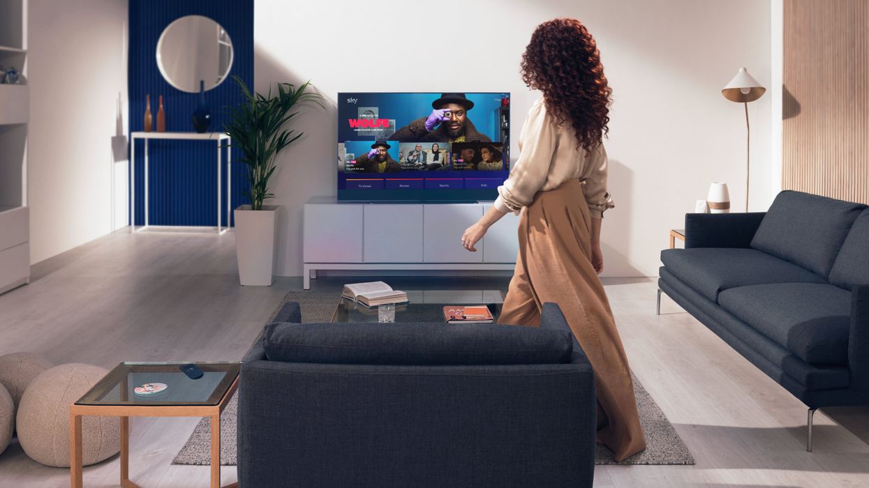a woman walks into a living room with a blue sky glass tv in the corner switched on showing the main entertainmentos menu