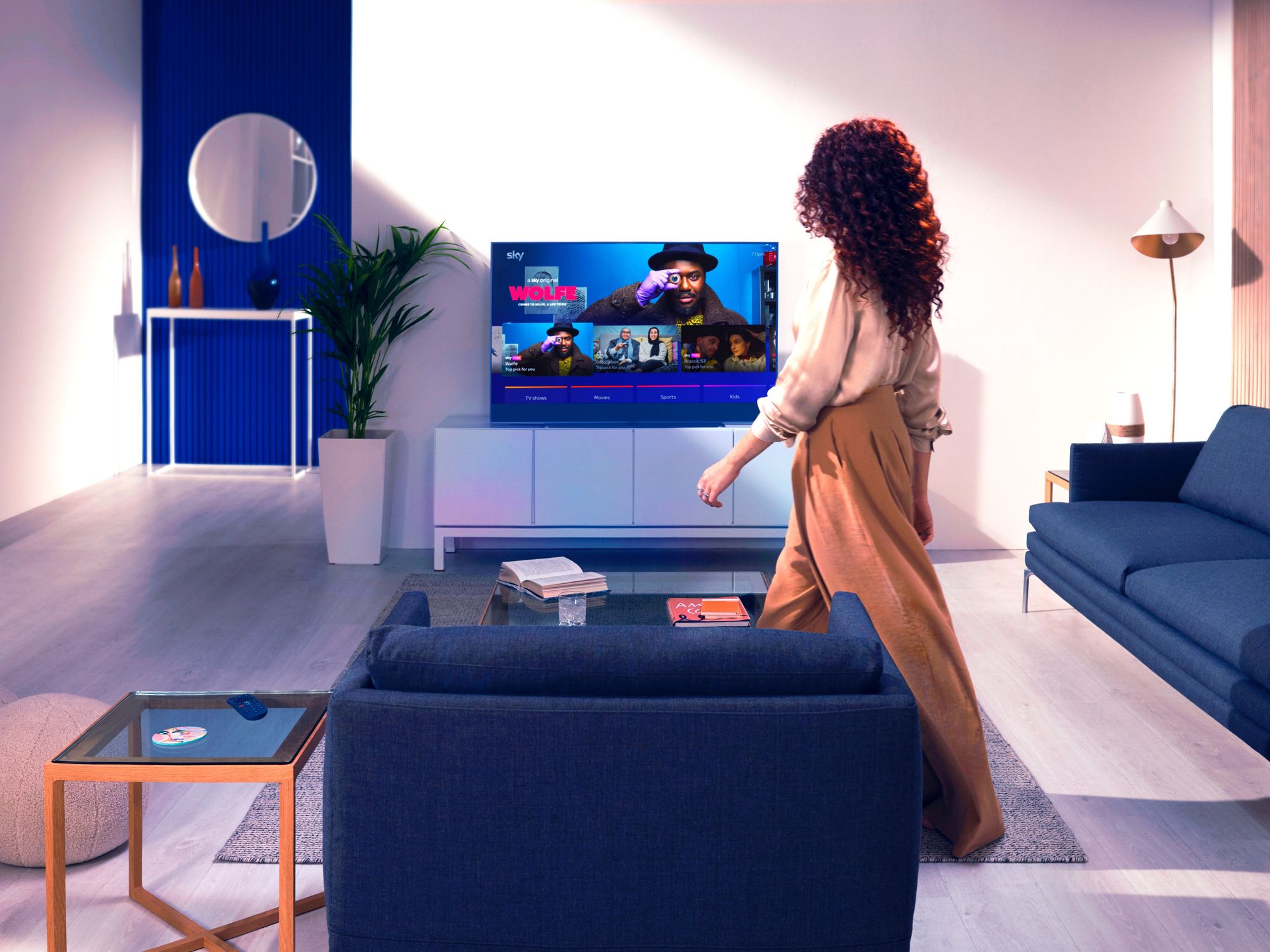 a woman walks into a living room with blue sofas and a sky glass television on a stand