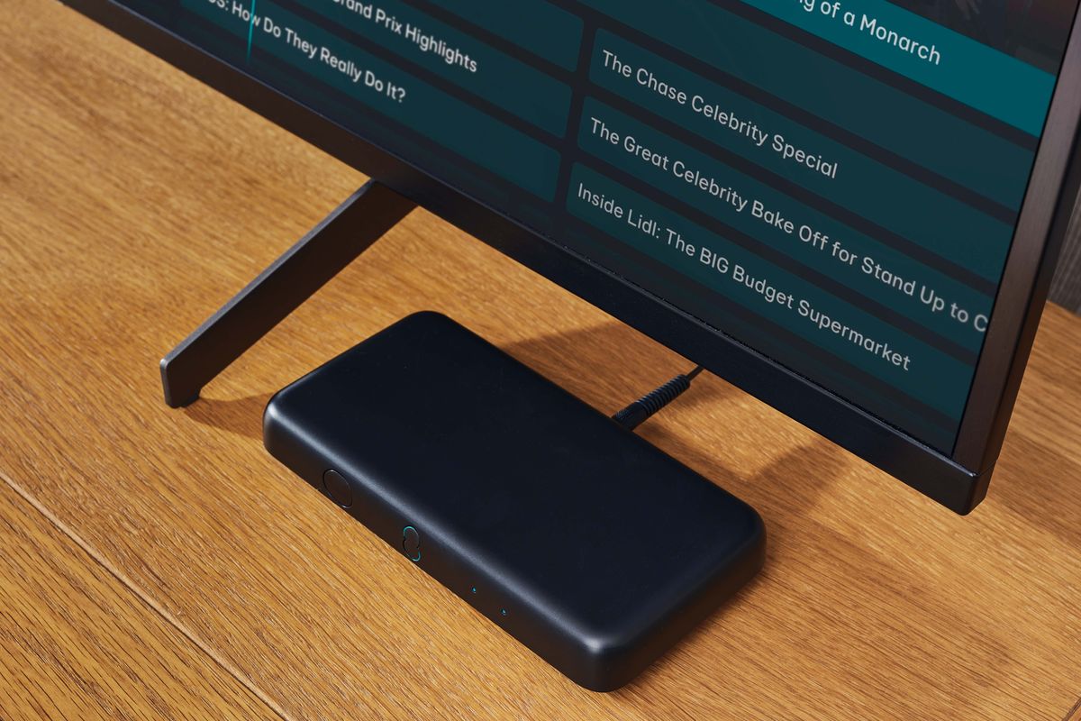 an ee tv box mini is shown on a wooden table, nestled beneath a flatscreen television with an EPG TV Guide shown on screen 