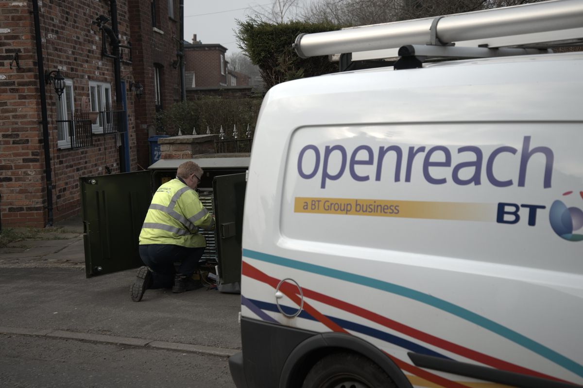 an openreach engineer is pictured making adjustments inside an exchange cabinet on the streets with a BT and openreach branded van parked beside him 