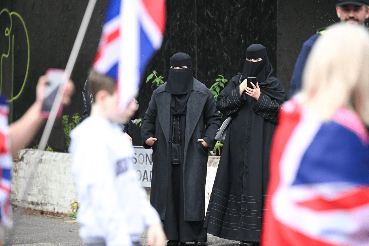 Anti-migrant protesters march past women wearing Burkas