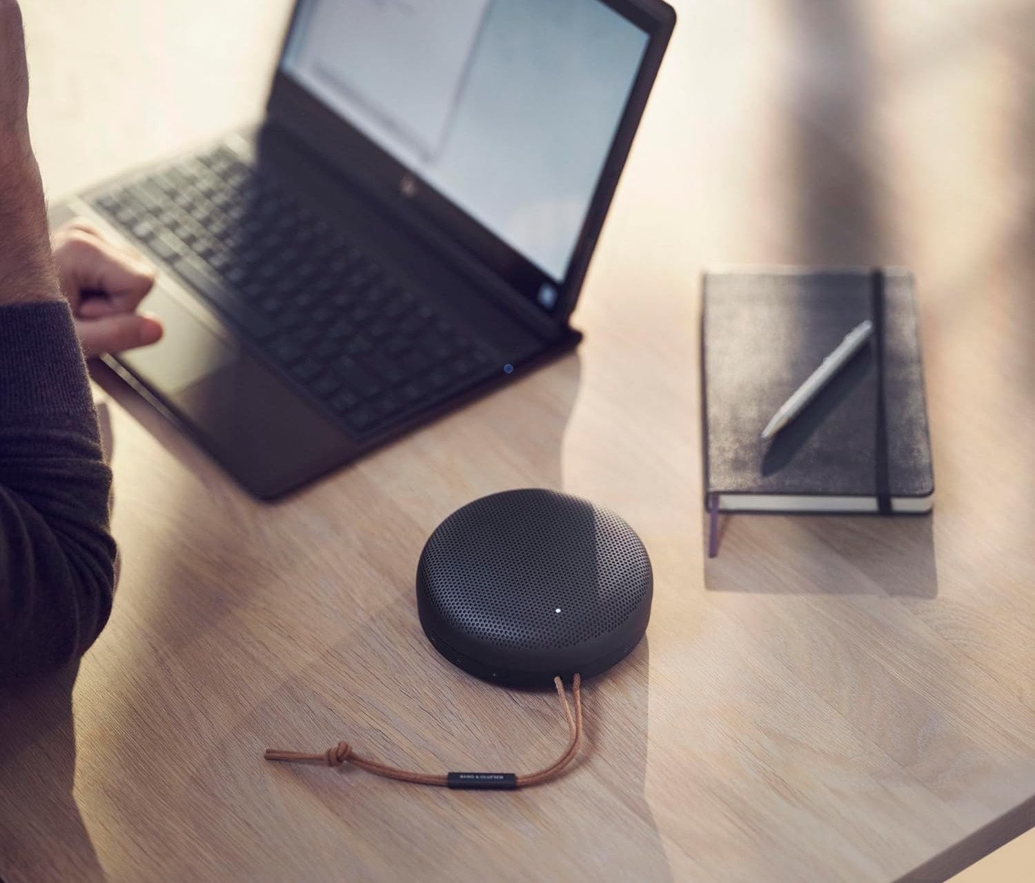 bang and olufsen bluetooth speaker on a desk beside someone working on a laptop
