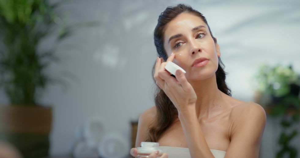 Brunette woman in a towel gently applying face cream under her eyes
