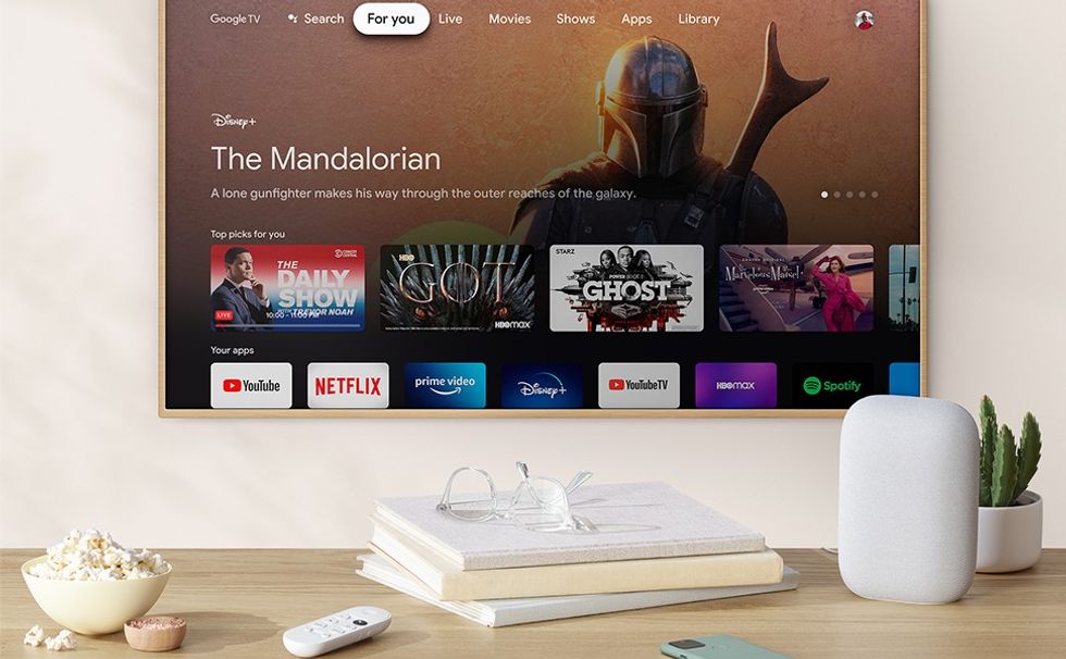 Chromecast with google tv on-screen with the remote lying on a table with books and google nest speaker beside it