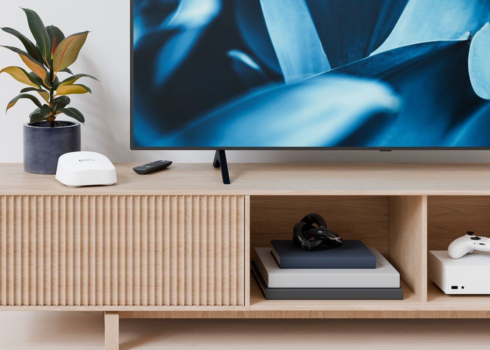 Eero 7 wireless router pictured on a table next to a television with a fire tv remote control and xbox series s