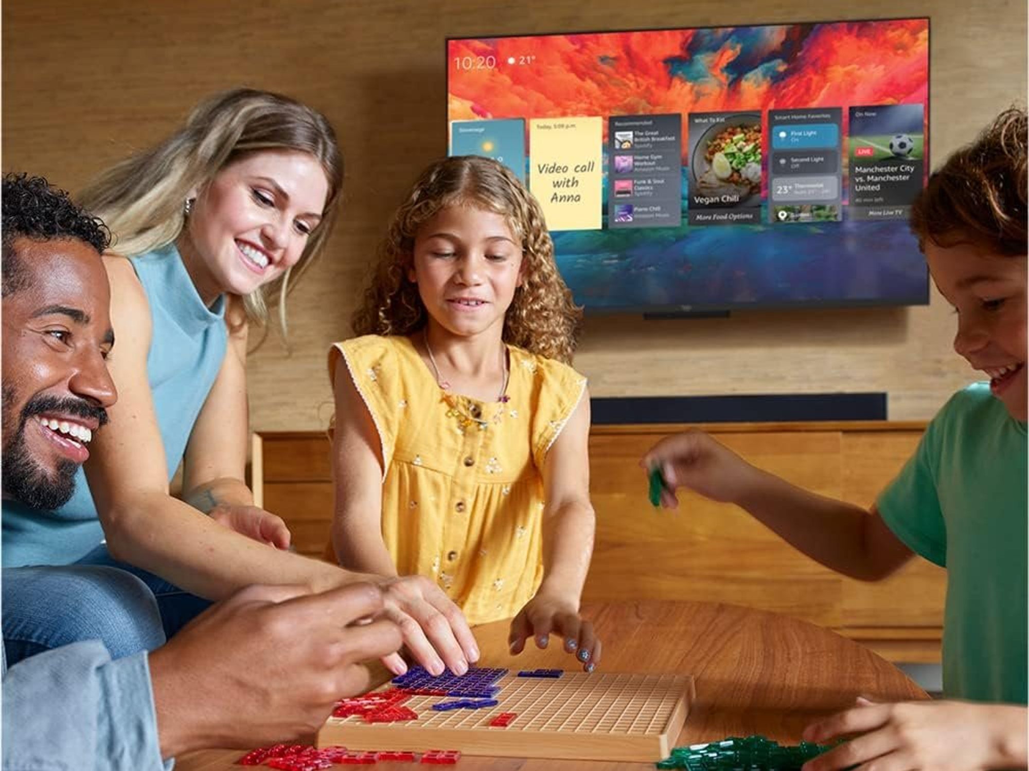 family playing a board game with the Fire TV Omni QLED in the background with Alexa widgets on-screen