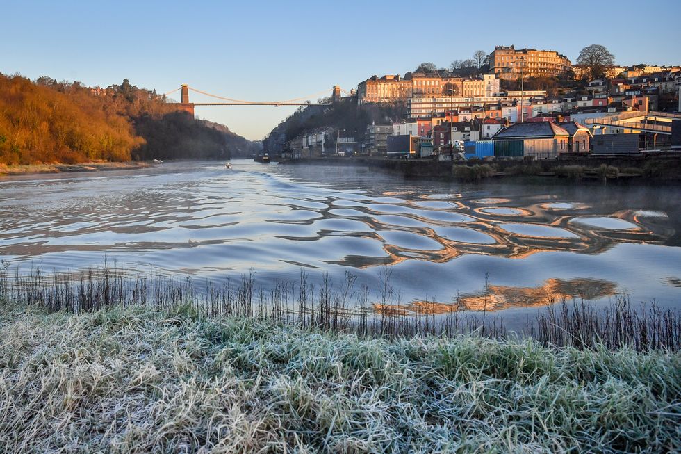 Frost forms on grass along the bank of the River Avon at Cumberland Basin in Bristol as the sun rises over Clifton Suspension Bridge.