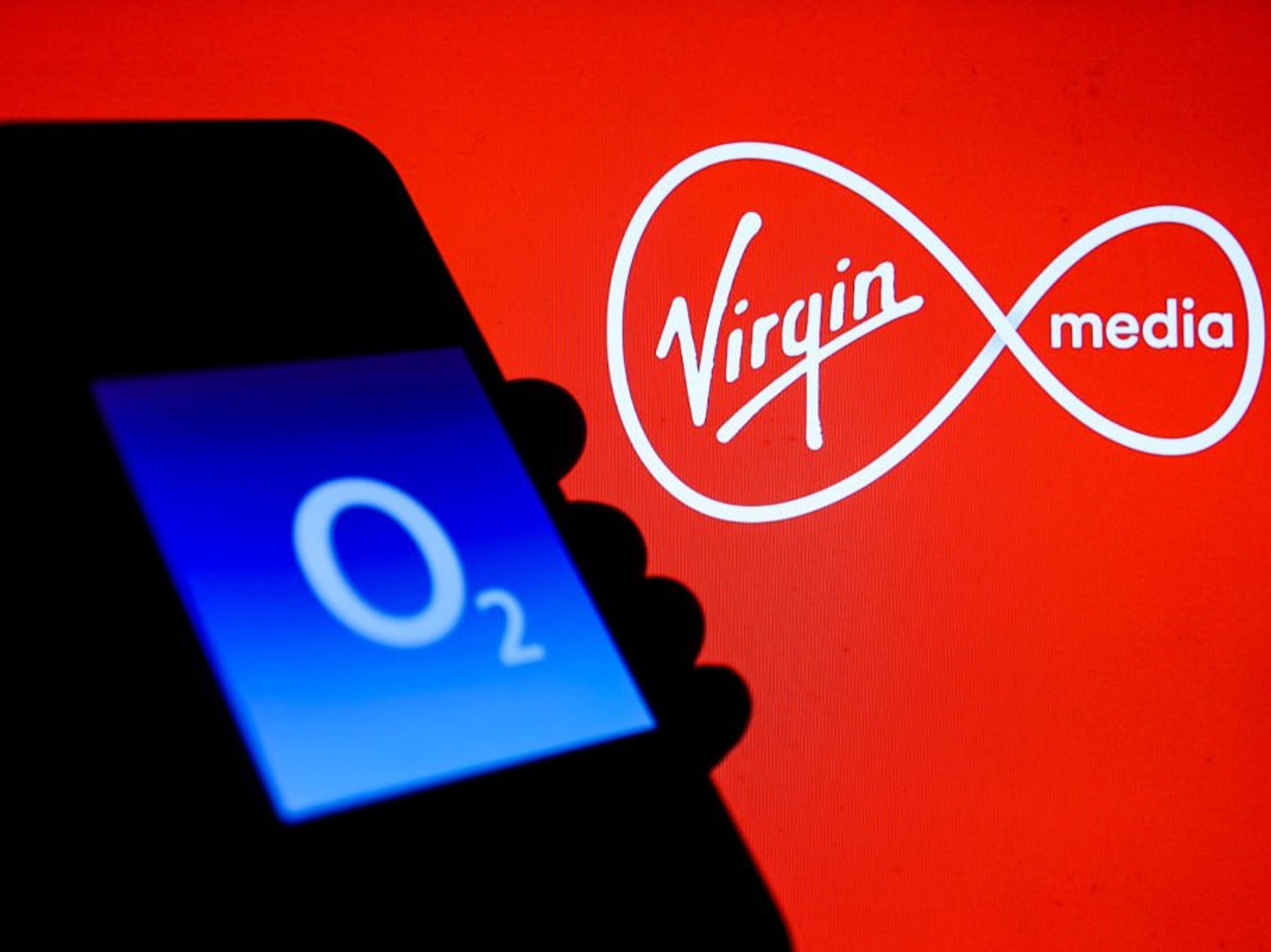 Hand holding phone with O2 logo displayed and Virgin Media logo displayed behind it
