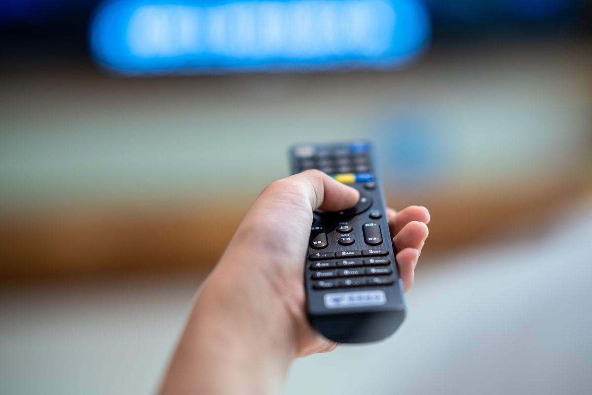 hand holds a remote control pointing at an out-of-focus smart tv in the background