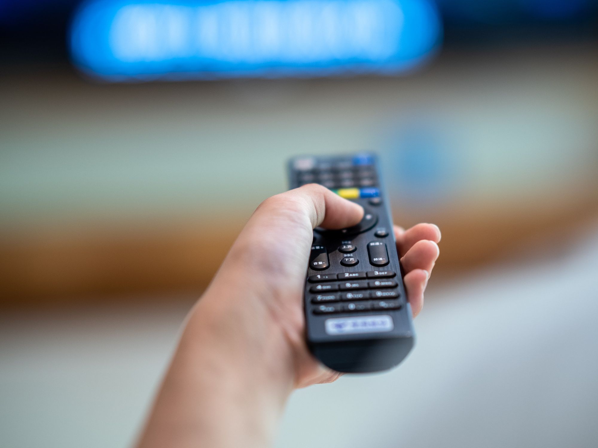 hand holds a remote control pointing at an out-of-focus smart tv in the background