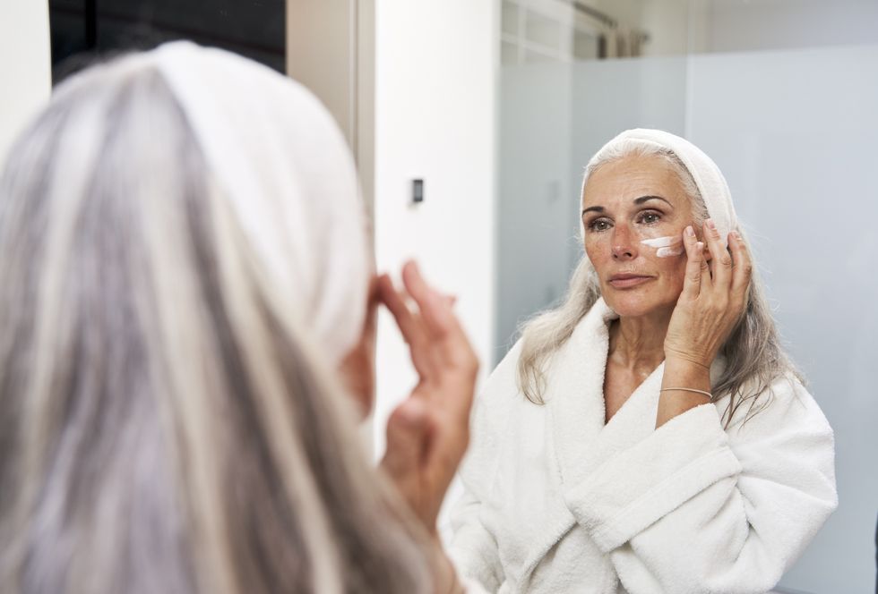 Mature woman with grey hair in a dressing robe and a hair band putting on face cream on her cheeks