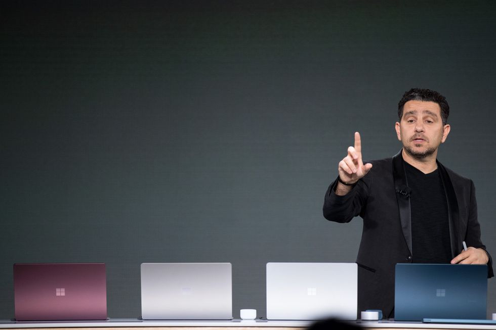 microsoft executive panos panay standing with four different coloured surface laptops on-stage during a presentation