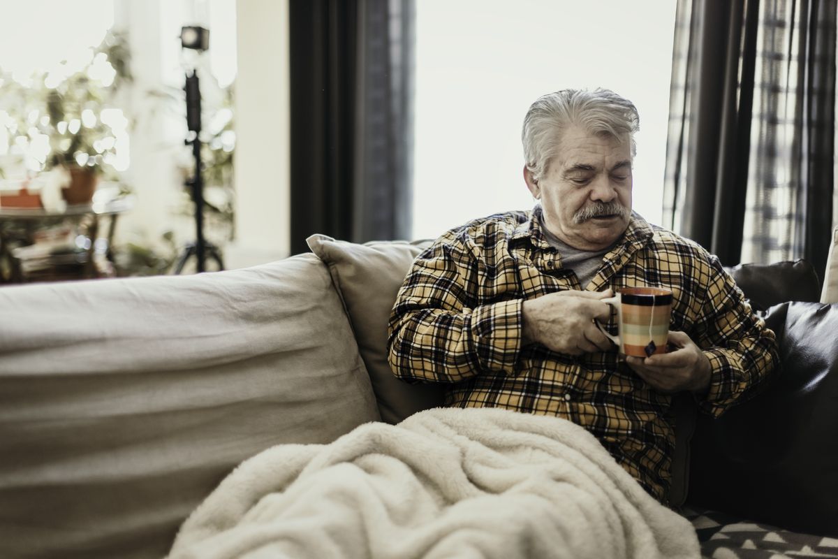 Pensioner holds mug and sits under blanket on sofa