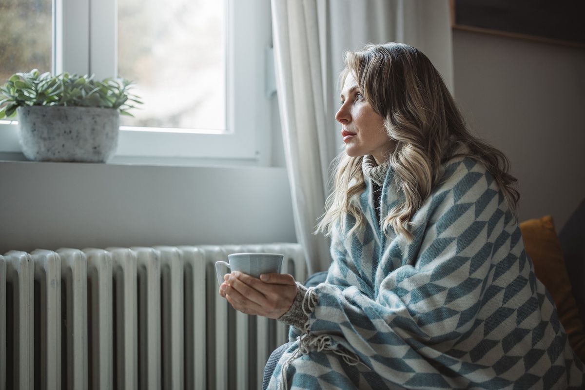 Person looks cold in blanket by radiator with mug in hands