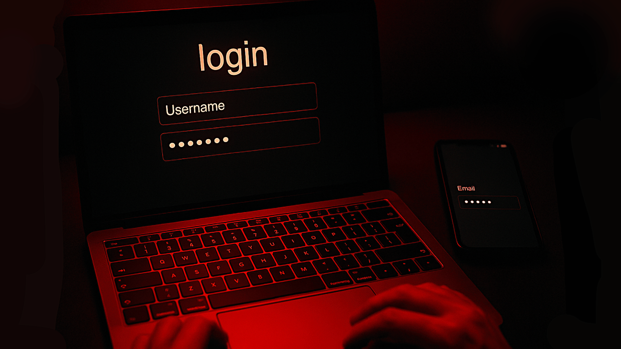 red lighting shines on hands on a laptop keyboard typing a password into a login screen with a smartphone with a password input required pictured to the side