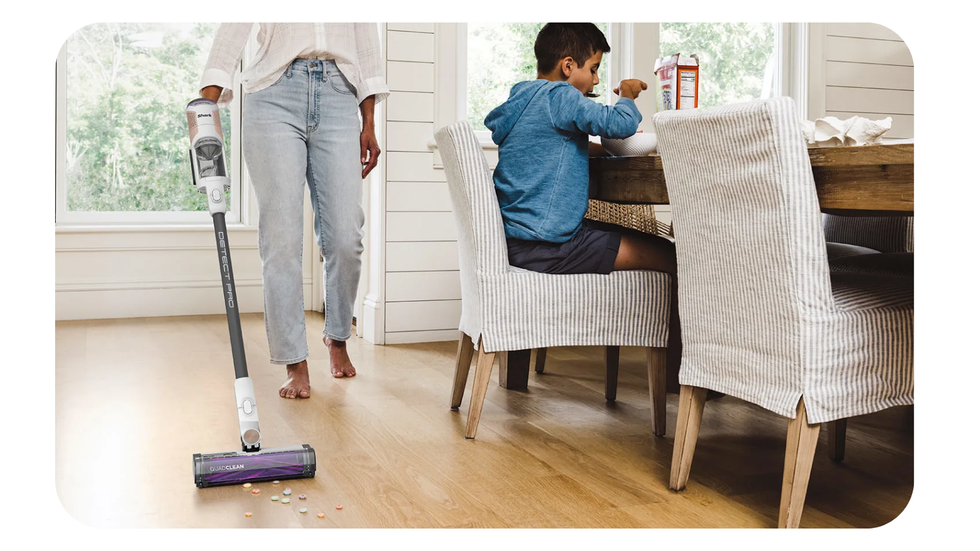 Shark Detect Pro cordless vacuum shown cleaning cereal from the floor while a child eats their breakfast at the table